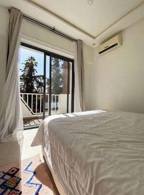 Une chambre avec un grand lit, une porte-fenêtre coulissante donnant sur un balcon et une bouche d’aération au plafond.