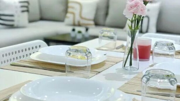Un comedor con una mesa blanca dispuesta para dos, un florero con flores rosadas y una pared de color amarillo brillante al fondo.