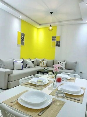 Un salon moderne avec un mur d’accent jaune, un canapé gris et un coin repas agrémenté d’assiettes et de couverts blancs.