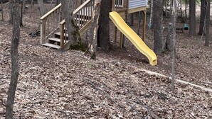 Une terrasse en bois avec un toboggan jaune, entourée d’arbres.