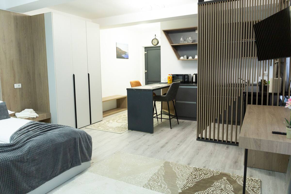 A modern hotel room with a bed, a desk, a chair, a kitchenette, and a wardrobe.