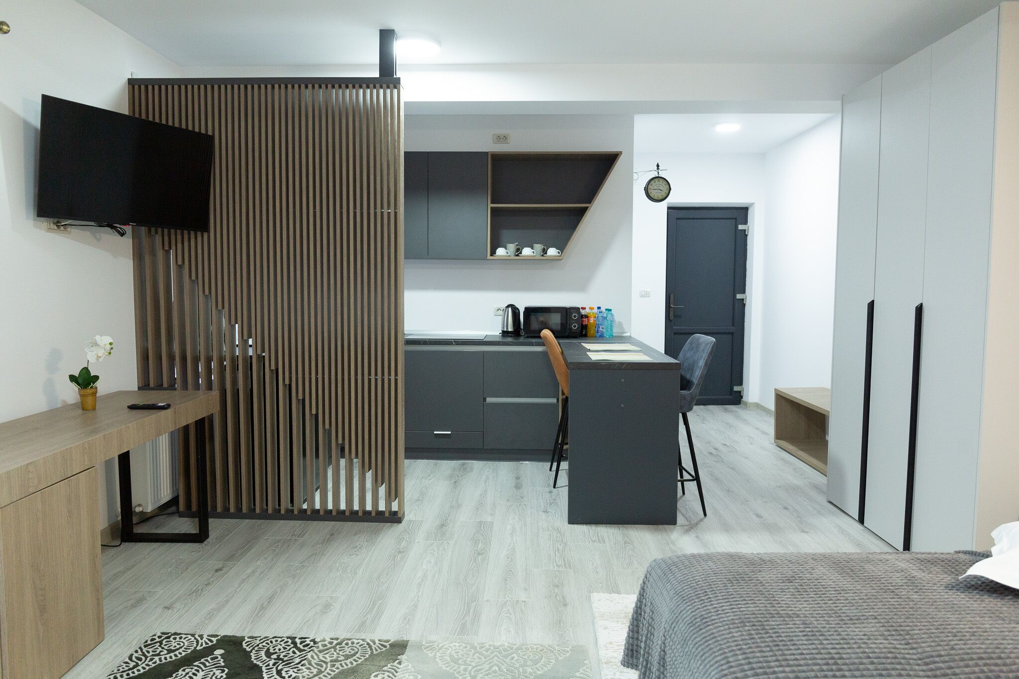 Ein modernes, kompaktes Hotelzimmer mit einer Küchenzeile, einem an der Wand montierten Fernseher, einer Holzwand und einem Bett mit einem gemusterten Bettüberwurf.