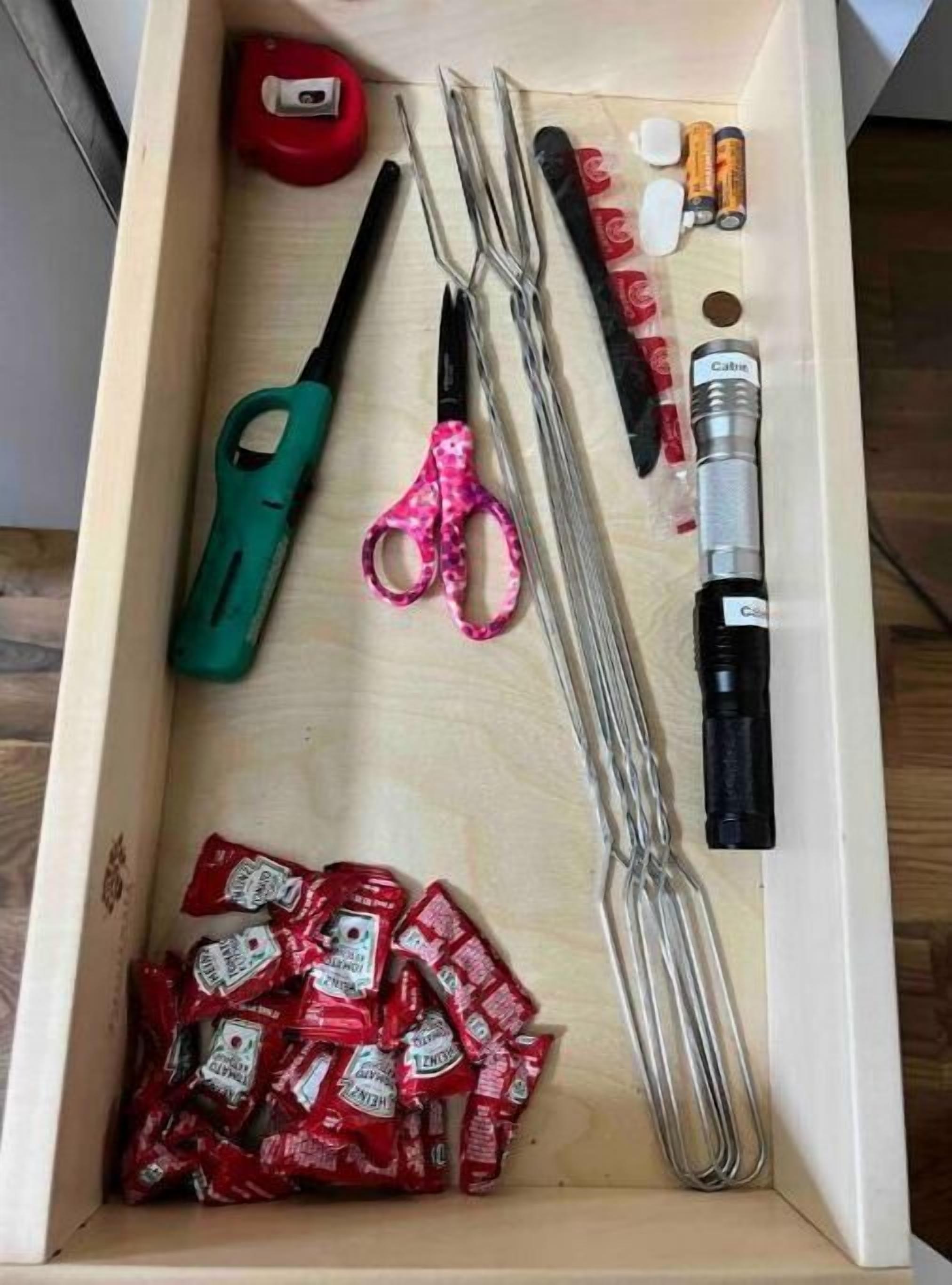 A drawer containing a pair of scissors, a tape measure, a lighter, a pen, and several packets of snacks.