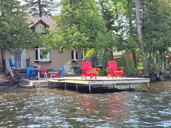 A dock with red chairs and a blue chair, a house with a dock, and trees in the background. - Rustic Pines Waterfront Cottage *CLEAR WATER* (Kawartha Lakes)