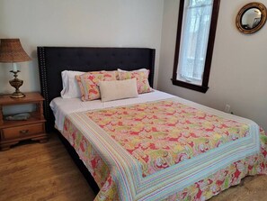A bedroom with a dark headboard, a colorful quilt, a bedside table with a lamp, and a window with a mirror.