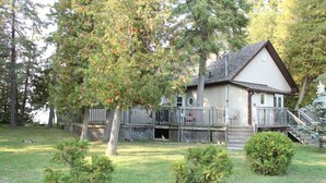 A two-story house with a deck, surrounded by trees and a grassy yard. - Rustic Pines Waterfront Cottage *CLEAR WATER* (Kawartha Lakes)