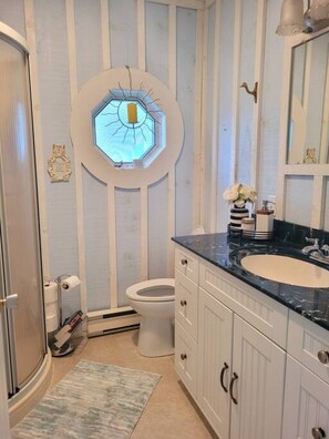 A bathroom with a white and blue striped wall, a round window with a sun design, a white toilet, a sink with a black countertop, and a mirror above the sink.