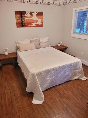 A bedroom with a large bed, two bedside tables, a window with blinds, and a wall-mounted painting.