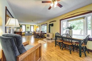 A spacious living room with a wooden floor, a ceiling fan, a dining area with a table and chairs, and large windows allowing natural light.
