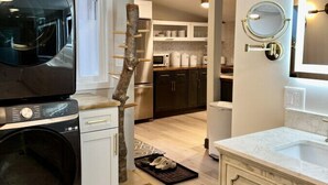 A modern kitchen with a built-in washer and dryer, a tree trunk as a decorative feature, and a well-lit bathroom with a mirror and sink.