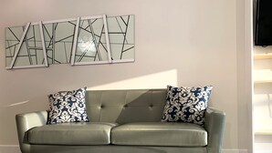 Ein graues Ledersofa mit Holzbeinen, zwei gemusterte Kissen und ein modernes abstraktes Kunstwerk darüber.