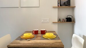 Ein Essbereich mit einem Holztisch, gedeckt für zwei Personen, zwei roten Tassen, zwei gelben Schalen und einem schwarzen Wasserkocher auf einem Regal.