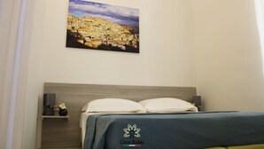 A hotel room with a bed, a nightstand, a lamp, and a painting of a town on the wall. - CASA DEGLI AMICI (Ruvo di Puglia)