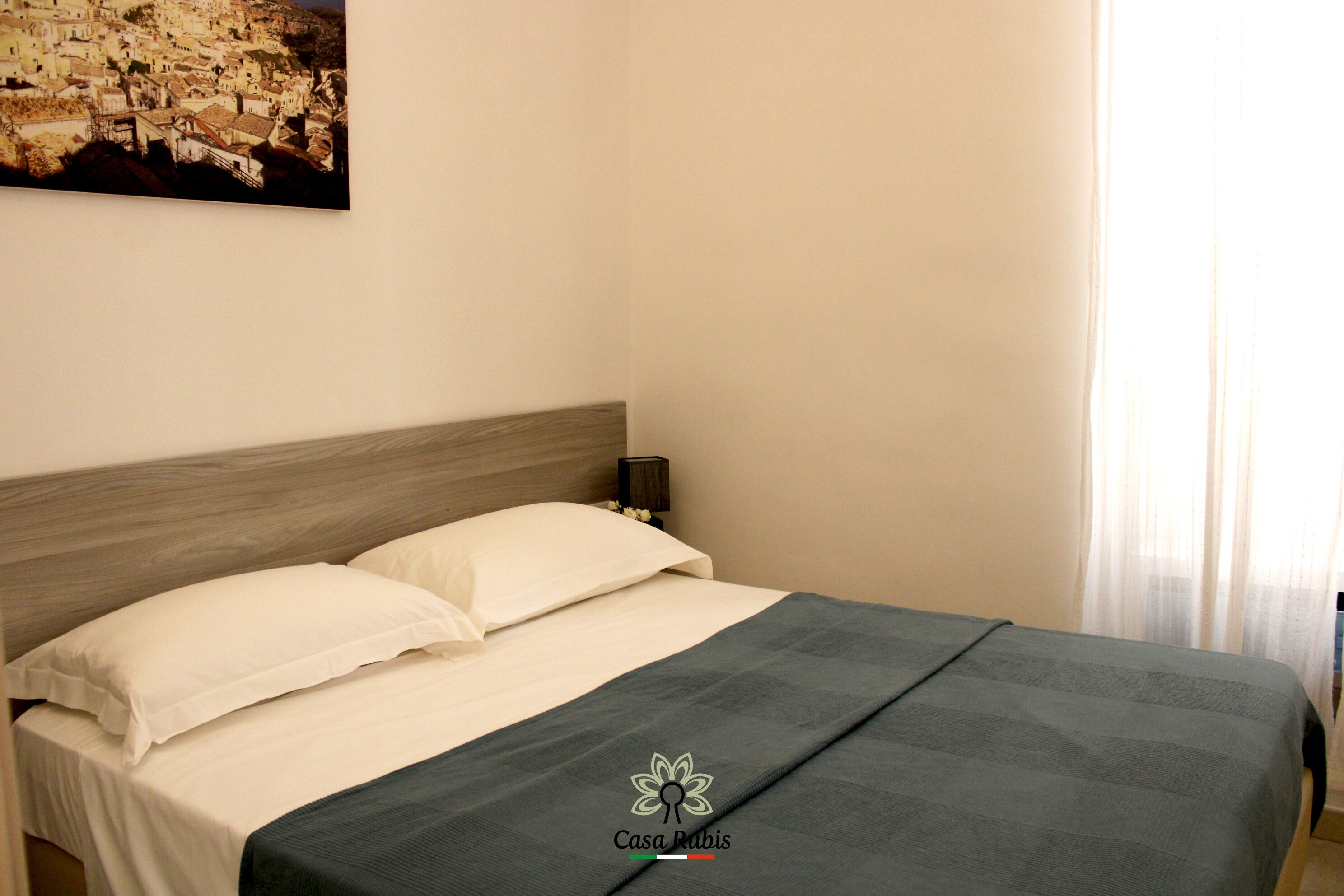 A neatly made bed with a wooden headboard, a nightstand with a lamp, and a painting of a town on the wall.