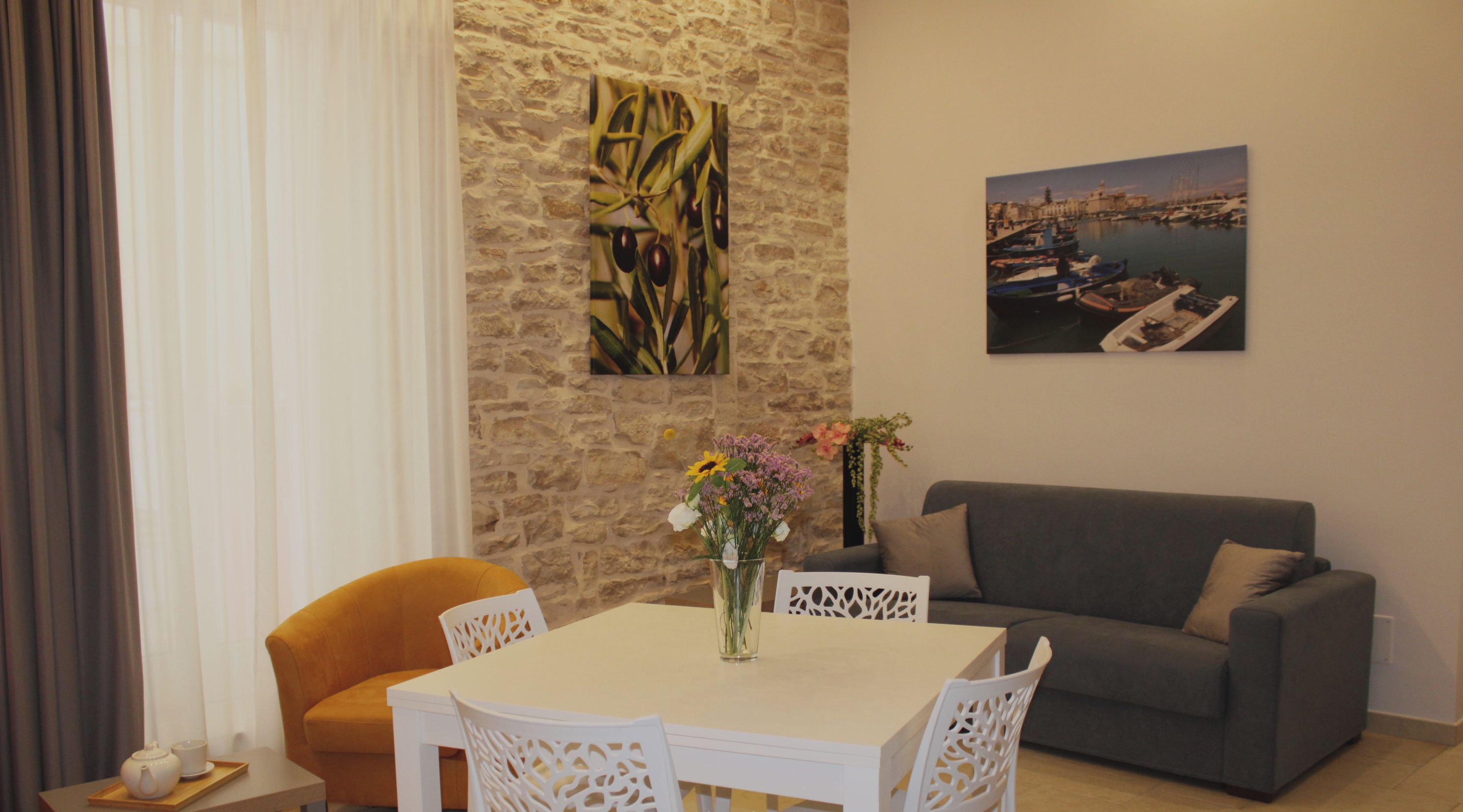 Uma sala de jantar com uma mesa e cadeiras brancas, um sofá cinzento e uma parede de pedra decorada com obras de arte.
