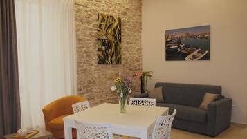 Uma sala de jantar com uma mesa e cadeiras brancas, um sofá cinzento e uma parede de pedra decorada com obras de arte.