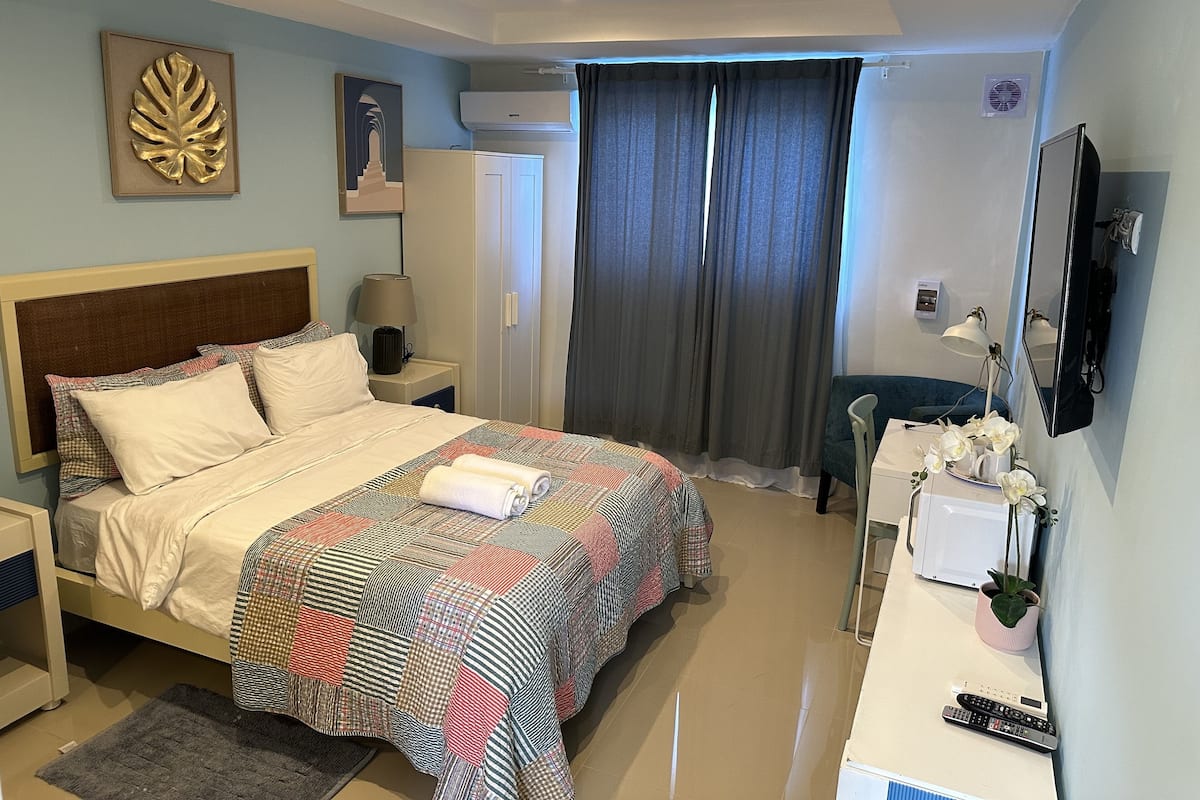 Habitación de hotel con cama, escritorio con silla, televisor, espejo, ventana con cortinas y aire acondicionado instalado en la pared.