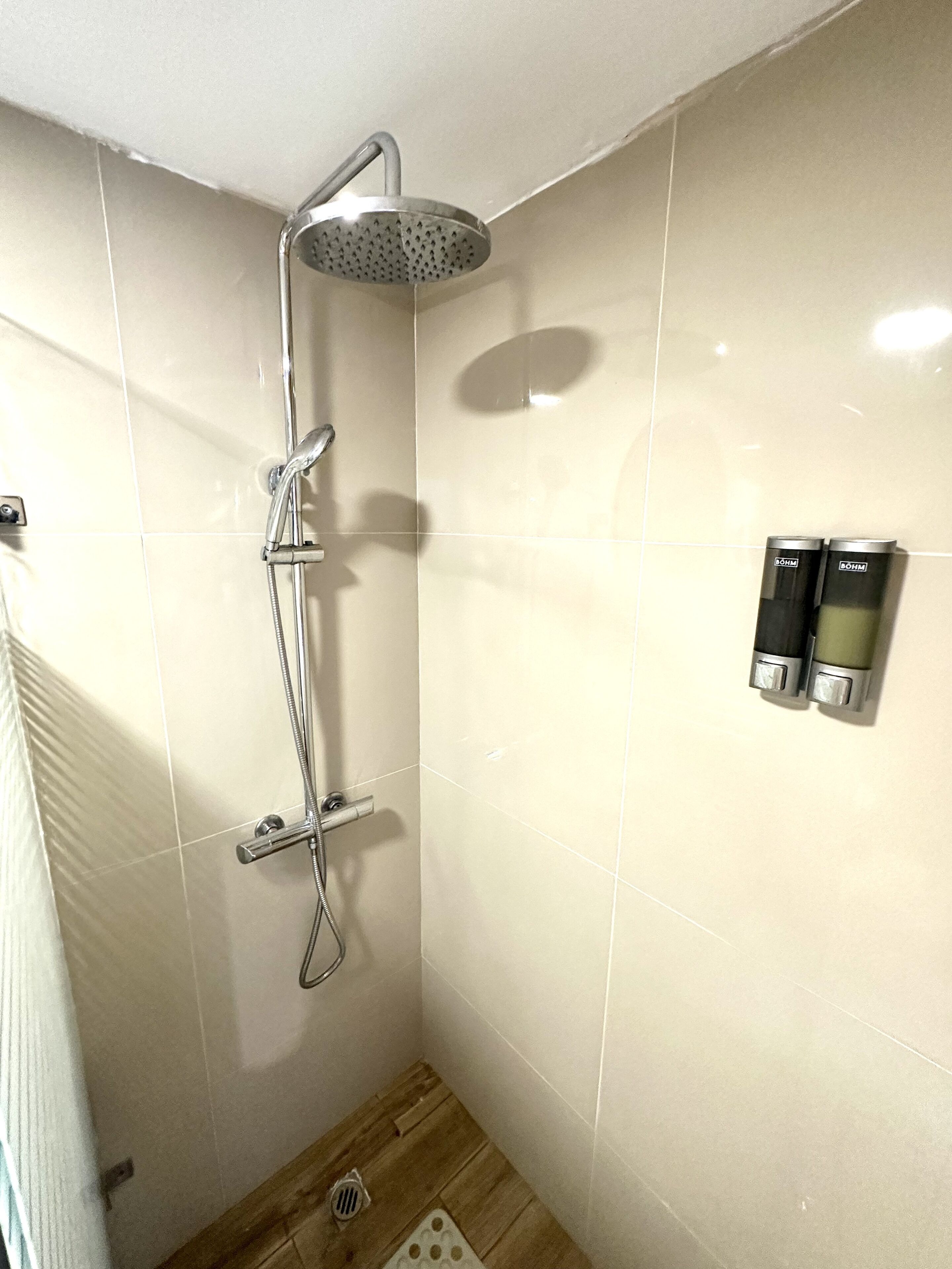 Pequeno espaço de chuveiro com azulejos, chuveiro manual, torneira na parede e dois dispensadores de sabonete.