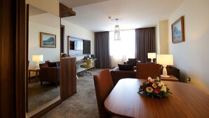 A hotel room with a wooden dining table, two armchairs, a sofa, a desk with a computer, a TV, and a large window with curtains. - Tuyap Palas (Istanbul)