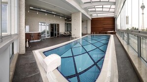 A large indoor swimming pool with a glass ceiling, a wooden wall, and a view of a tower. - Tuyap Palas (Istanbul)