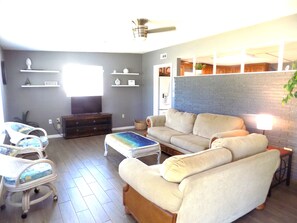 Ein Wohnzimmer mit einer Couch, einem Fernseher, einem Couchtisch und einem Sessel. Regale mit Dekoration und einer Pflanze sind ebenfalls vorhanden.