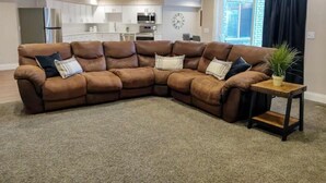 A L-shaped sectional sofa with multiple cushions, a small wooden side table with a plant, and a kitchen area in the background. - Cheery and Bright four Bedroom Walkout Basement (Highland)