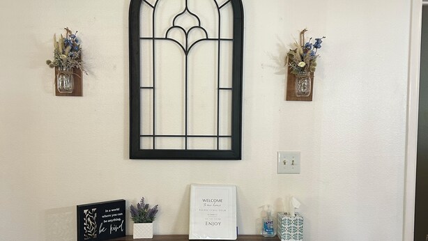 A wall-mounted window with a black frame, flanked by two small wall-mounted planters with dried flowers. Below, a wooden shelf holds a framed inspirational quote, a small potted plant, and a tissue box. - Sunny Days! Room for everyone and Pet-Friendly! (Pocatello)