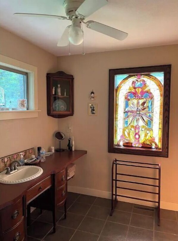Un baño con tocador de madera, ventilador de techo, ventana con vitrales y un taburete pequeño.