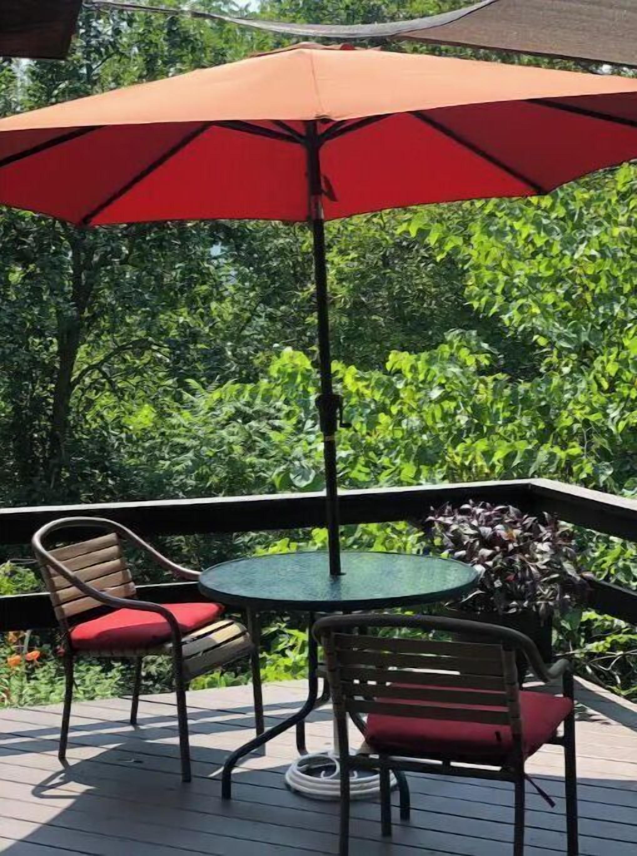 Une terrasse extérieure avec une table ronde et deux chaises, un parasol rouge et une végétation luxuriante en arrière-plan.