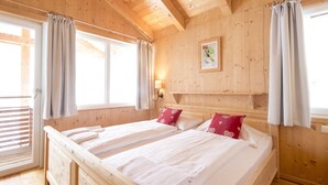 A wooden room with a large bed, two pillows, a wall-mounted light, and a window with curtains. - Chalet #7 with IR-sauna (Turrach)