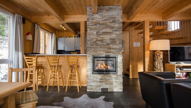A cozy cabin interior with a stone fireplace, wooden furniture, and a kitchen area. - Chalet #7 with IR-sauna (Turrach)