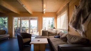 Ein gemütliches Holzinterieur mit einer Couch, Sesseln und einem Couchtisch. Große Fenster bieten einen Blick ins Freie.