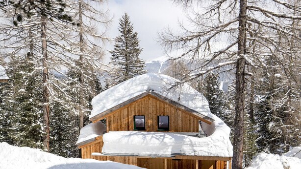 Eine Holzhütte mit schneebedecktem Dach, umgeben von hohen Kiefern in einer verschneiten Landschaft.