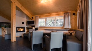 En mysig stuga med en öppen spis, en matplats och en sittgrupp med utsikt över trädtopparna genom fönstren.