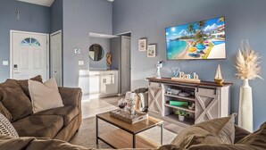 Ein Wohnzimmer mit braunem Sofa, einem Couchtisch mit Tablett, einem Kamin mit der Aufschrift 'Beach' und einem Fernseher, der eine Strandansicht zeigt.