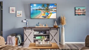 Ein Wohnzimmer mit an der Wand montiertem Fernseher, einem Holztisch und einem Bücherregal mit verschiedenen Gegenständen.