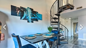 Un espace repas avec une table en marbre noir, des chaises recouvertes d’un tissu bleu et un escalier en colimaçon.