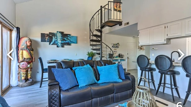 Un salon moderne avec un canapé en cuir noir, des coussins bleus, un escalier en colimaçon et un coin cuisine doté de meubles blancs.