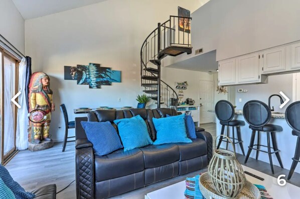 A modern living room with a black leather sofa, blue cushions, a spiral staircase, and a kitchen area with white cabinets.