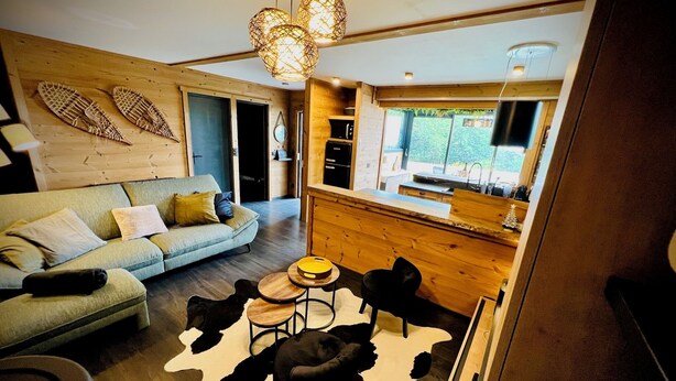 Ein modernes Wohnzimmer mit einer Couch, einem Couchtisch und einem Teppich. Durch einen offenen Durchgang ist eine Küchenzeile erkennbar.