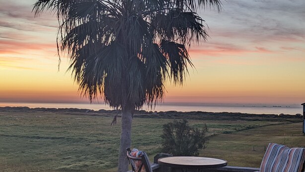 Une terrasse avec une table ronde et deux chaises à rayures, donnant sur un champ verdoyant et sur un coucher de soleil sur l’eau.