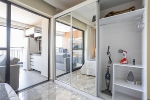 Une chambre d’hôtel moderne dotée d’une porte-fenêtre coulissante donnant sur un balcon, d’une petite kitchenette, d’un lit avec une couette blanche et d’une étagère où se trouvent un aspirateur et un fer à repasser.
