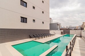 A rooftop swimming pool with lounge chairs, a tiled wall, and a cityscape view.