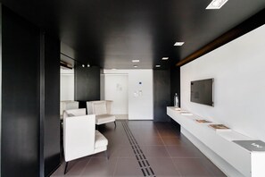 A modern reception area with a white reception desk, a flat-screen TV, and a seating area with white chairs. - Vita Bom Retiro (São Paulo)
