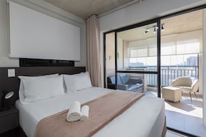 A hotel room with a bed, a desk with a lamp, a chair, a balcony with a view, and a sliding glass door.