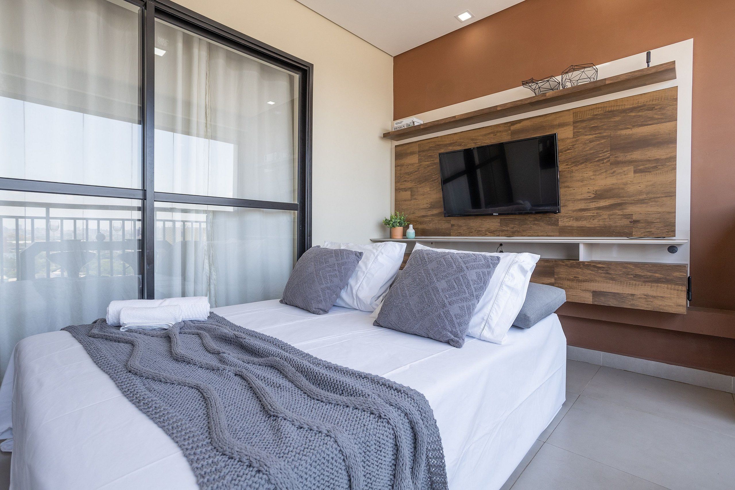 Um quarto moderno com uma cama grande, televisão fixada em uma parede de madeira e uma janela com cortinas.