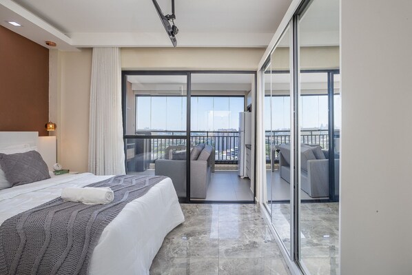 A modern hotel room with a large bed, a balcony with seating, and a city view.