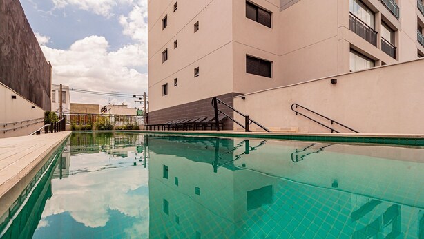 A swimming pool with clear water, surrounded by buildings with balconies and a wooden deck. - Vita Bom Retiro (São Paulo)