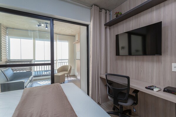A hotel room with a bed, a desk with a chair, a TV, and a balcony with a chair and a table.