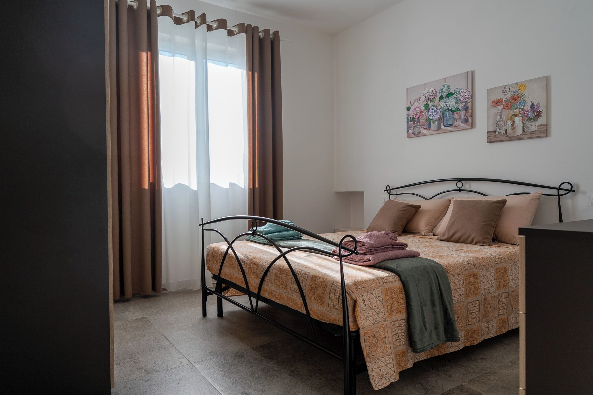 A bedroom with a metal bed frame, a patterned bedspread, and a green throw blanket. There are two paintings on the wall above the bed.